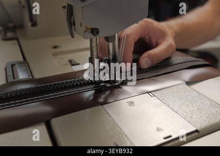 Worker operating a heavy duty sewing machine, stitching a durable black zipper onto a brown waterproof fabric, representing craftsmanship and industri Stock Photo
