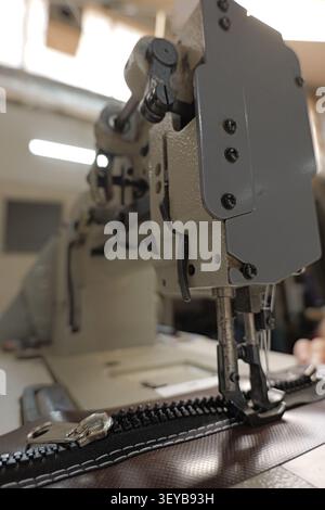 Industrial sewing machine mechanism performing precision stitching, automatically attaching a long black zipper onto a brown textile product, showcasi Stock Photo