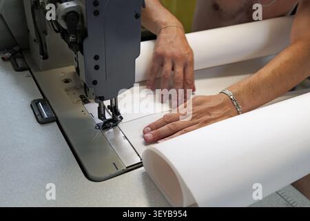 Worker hands guide plain white fabric under the needle of an industrial sewing machine, performing a precise task in a manufacturing process Stock Photo