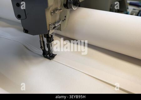 Industrial sewing machine performing stitching on a long roll of white fabric, highlighting the automated precision in textile production and the manu Stock Photo