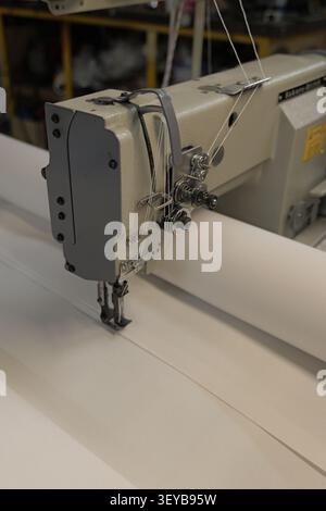 Industrial sewing machine performing a close up stitch on a white fabric curtain in a textile factory, creating durable seams for various manufacturin Stock Photo