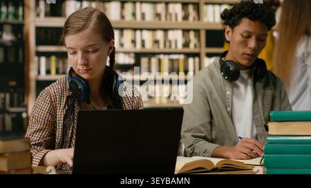 Young woman student reading and working on laptop between open books at library table with shelves behind her for digital learning and focused academic research. Stock Photo
