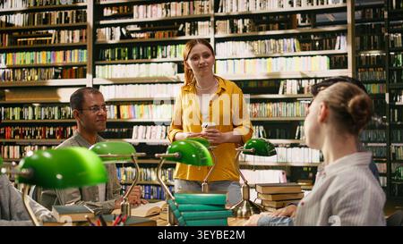 Tutor in yellow speaks to university students gathered at library table with green lamps, using books and laptop during group study session. Collaborative learning and guidance for classmates. Stock Photo