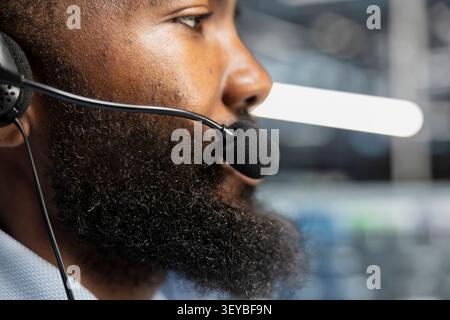 Server facility staff member in remote call providing customer support, troubleshooting damages. African American man in server facility talking with customers, working with microphone. Stock Photo