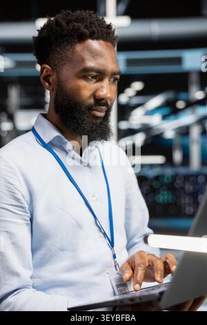 African American computer scientist uses a laptop in a server room to manage data backups, resolve errors and support recovery. Stock Photo