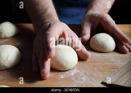 Hands shaping smooth dough balls on a floured wooden board before proofing. Preparation of homemade burger buns or bread rolls in a home kitchen. Stock Photo