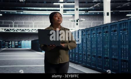 Cheerful systems engineer walking in server facility with notebook computer, checking hardware for system stability. Stock Photo