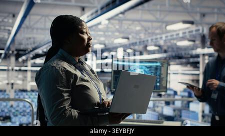 Server facility supervisor on industrial platform working to improve AI systems processing speed. African American server facility supervisor supervising equipment supporting AI workloads. Stock Photo