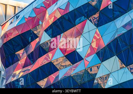 Striking architectural facade composed of triangular panels, reflecting vibrant colors and urban environment, showcasing dynamic geometric patterns wi Stock Photo