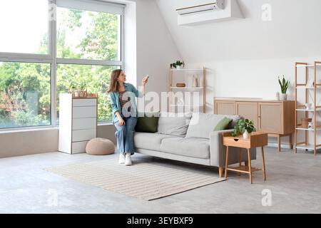 Young woman using remote control to operate air conditioner in living room Stock Photo