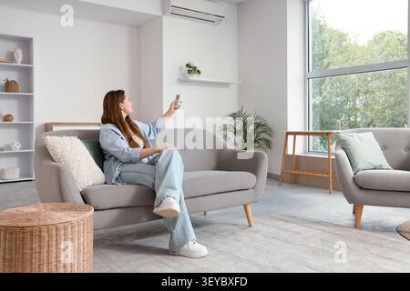 Woman sitting on sofa using remote to control air conditioner Stock Photo
