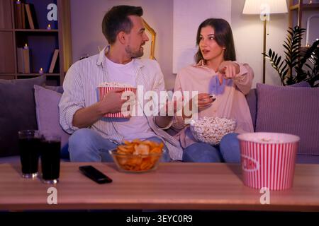 Shocked couple with potato chips and popcorn watching TV at home in evening Stock Photo