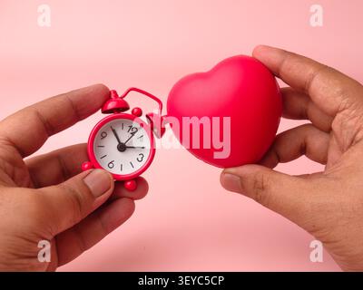 Two hands hold a small red mechanical alarm clock next to a matching red stress ball heart over pink. Stock Photo