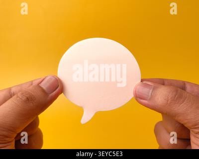 Two hands hold a blank white speech bubble cutout horizontally against a bright yellow studio background. Stock Photo
