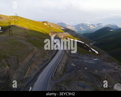 High angle perspective of an empty asphalt road running along a green grassy ridge under a dramatic cloudy sky with mountains Stock Photo