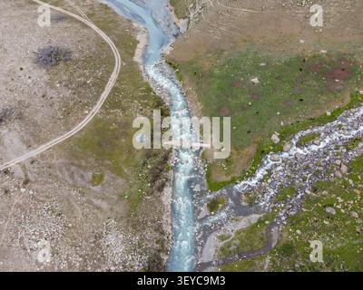 High angle overhead drone shot looking down at a small wooden bridge crossing a turquoise mountain river stream on a grassy plain Stock Photo
