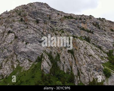 High angle perspective focusing on the massive steep gray limestone cliff face of a grand mountain with scattered small trees Stock Photo