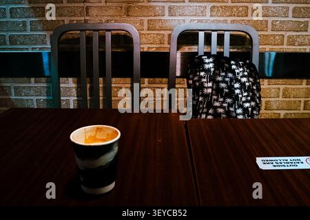 Paper Coffee Cup on Cafe table beside empty chairs and a backpack, capturing a quiet moment in a modern coffee shop. Stock Photo