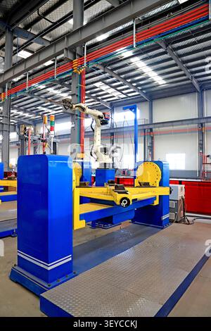 This photograph captures a state-of-the-art robotic welding workstation in a spacious, high-ceilinged industrial factory. A white articulated robotic Stock Photo