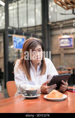 Focused lifestyle portrait of a Southeast Asian Isan transgender businesswoman in her 40s looking down at tablet while working at a cafe table. This v Stock Photo