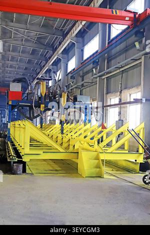 A dramatic vertical view of a heavy-duty automated welding machine positioned above a long, bright yellow steel structural frame assembly line in a sp Stock Photo