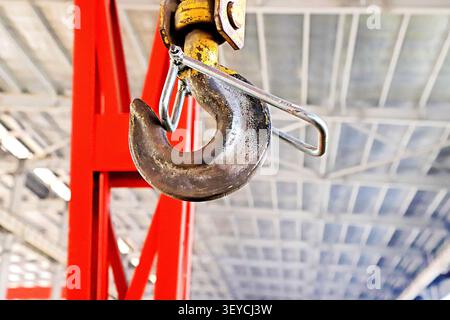 A dramatic low-angle close-up of a heavily rusted industrial crane hook, suspended from a bright red gantry crane in a spacious factory workshop. The Stock Photo