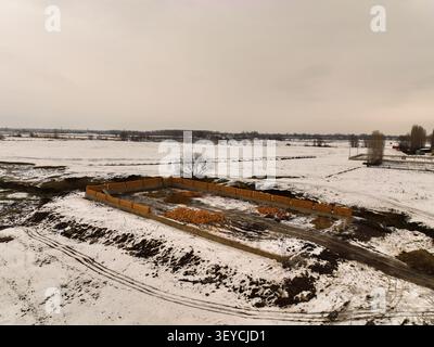 Wide aerial view of a construction foundation and stacks of bricks in a vast, snow-covered rural field under an overcast sky Stock Photo