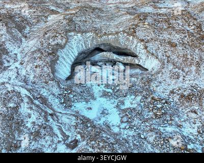 A top-down aerial view providing a detailed look at the archway and opening of a small ice cave or crevasse in the glacier's rocky moraine Stock Photo
