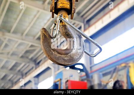 A dramatic, vertically oriented close-up of a heavily rusted crane hook, suspended from an overhead lifting system in a factory workshop. The weathere Stock Photo