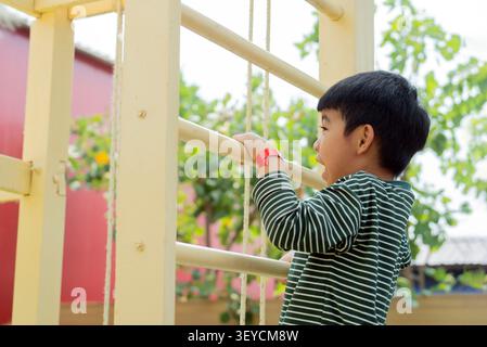 Kid exercise for health and sport concept. Side view of happy Asian child boy playing and climbing to a steel bar at the playground. Stock Photo