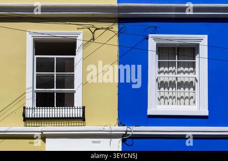 Colorful building facade with yellow and blue walls, two white framed windows, and overhead utility wires in bright sunlight. Bold urban architecture Stock Photo