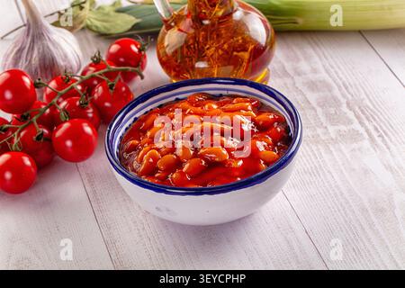Bowl of baked beans in tomato sauce surrounded by fresh cherry tomatoes, garlic, celery on rustic white wood. Warm lighting highlights reds oranges. F Stock Photo