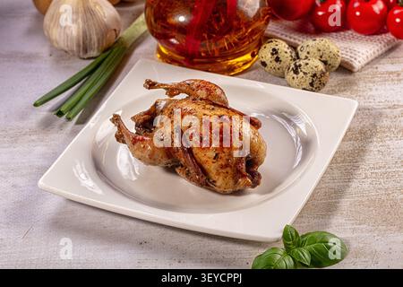 Golden brown roasted quail on white plate. Fresh garlic, green onions, cherry tomatoes, quail eggs, basil, infused oil in background Stock Photo