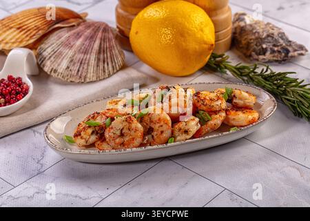 Plated grilled shrimp garnished with green herbs beside whole lemon rosemary sprigs seashells on marble surface. Warm lighting highlights golden shrim Stock Photo