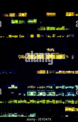 Illuminated office windows on a dark glass skyscraper facade at night, creating an abstract pattern of light and shadow. Stock Photo
