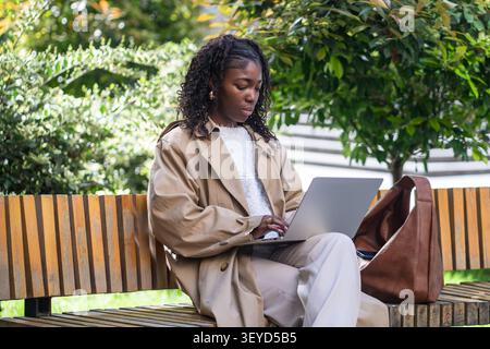 Focused african americam businesswoman sits on bench in park working online on laptop on day break Stock Photo
