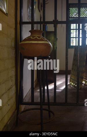A classic Sri Lankan terracotta water pitcher (Kalaya) displayed on a tall metal stand against a rustic interior wall with natural window lighting. Stock Photo
