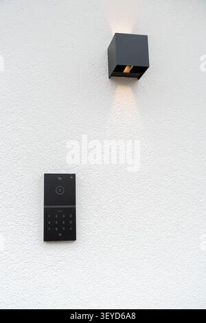 A stylish black smart doorbell with a keypad and camera mounted on a white textured wall below a modern black wall lamp emitting soft light. Ideal for Stock Photo