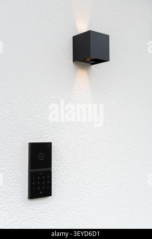 A stylish black smart doorbell with a keypad and camera mounted on a white textured wall below a modern black wall lamp emitting soft light. Ideal for Stock Photo
