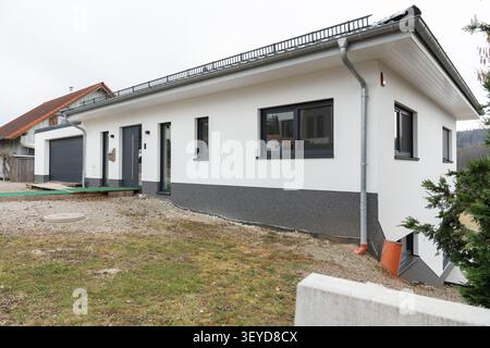 Modern single-story house with a crisp white exterior and contrasting dark windows, situated on a gently sloped yard. The minimalistic design Stock Photo