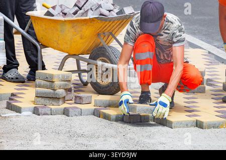 A group of workers is placing bricks to create a new pavement on a busy city street. Some bricks are stacked nearby, while one worker is on the ground Stock Photo
