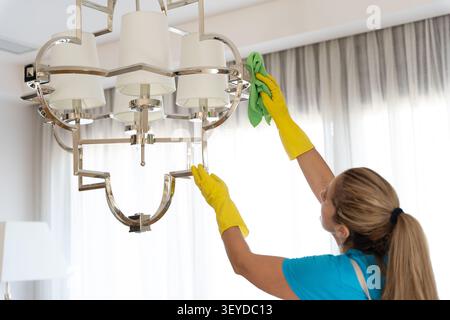 Woman in yellow gloves dusting a ceiling light fixture with a green cloth Stock Photo