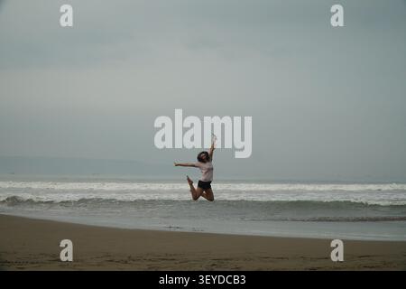 Full length view of a woman jumping in mid-air on a sandy beach with ocean waves under overcast sky in Indonesia Stock Photo