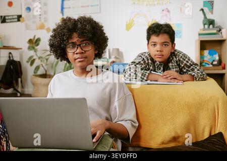 Portrait of Black Teenager Using Laptop with Child Studying at Home Stock Photo