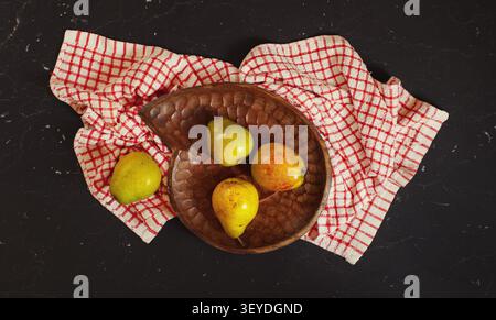 Top down view, green and yellow pears in wooden carved bowl, red chequered tablecloth and black marble board under Stock Photo