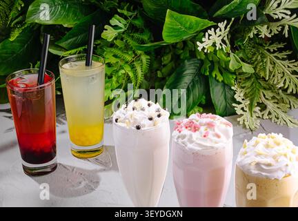Different sweet beverages stand on modern restaurant table. Refreshing summer cocktails are served against green leaves. Stock Photo