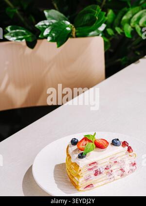 A beautiful slice of layered dessert is placed delicately on a white plate. Fresh strawberries and blueberries top the dessert, adding a vibrant touch. Green leaves create a natural backdrop. Stock Photo