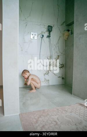 Baby playing in shower, in shower stall with marble tiles, with showerhead, bathtub mat, with marble tile, metal showerhead texture, from a side view Stock Photo