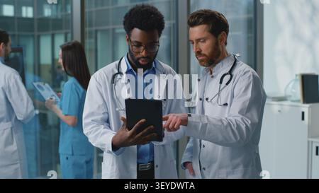 Two multiethnic doctors men pensive discussing medicine test results looking in digital tablet in clinic. African American therapist and Caucasian Stock Photo