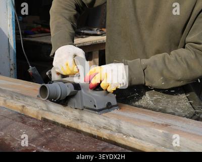 A craftsman wearing gloves and a khaki-colored top is planing a wooden beam with a grey electric planer. Fine shavings fly from the tool Stock Photo